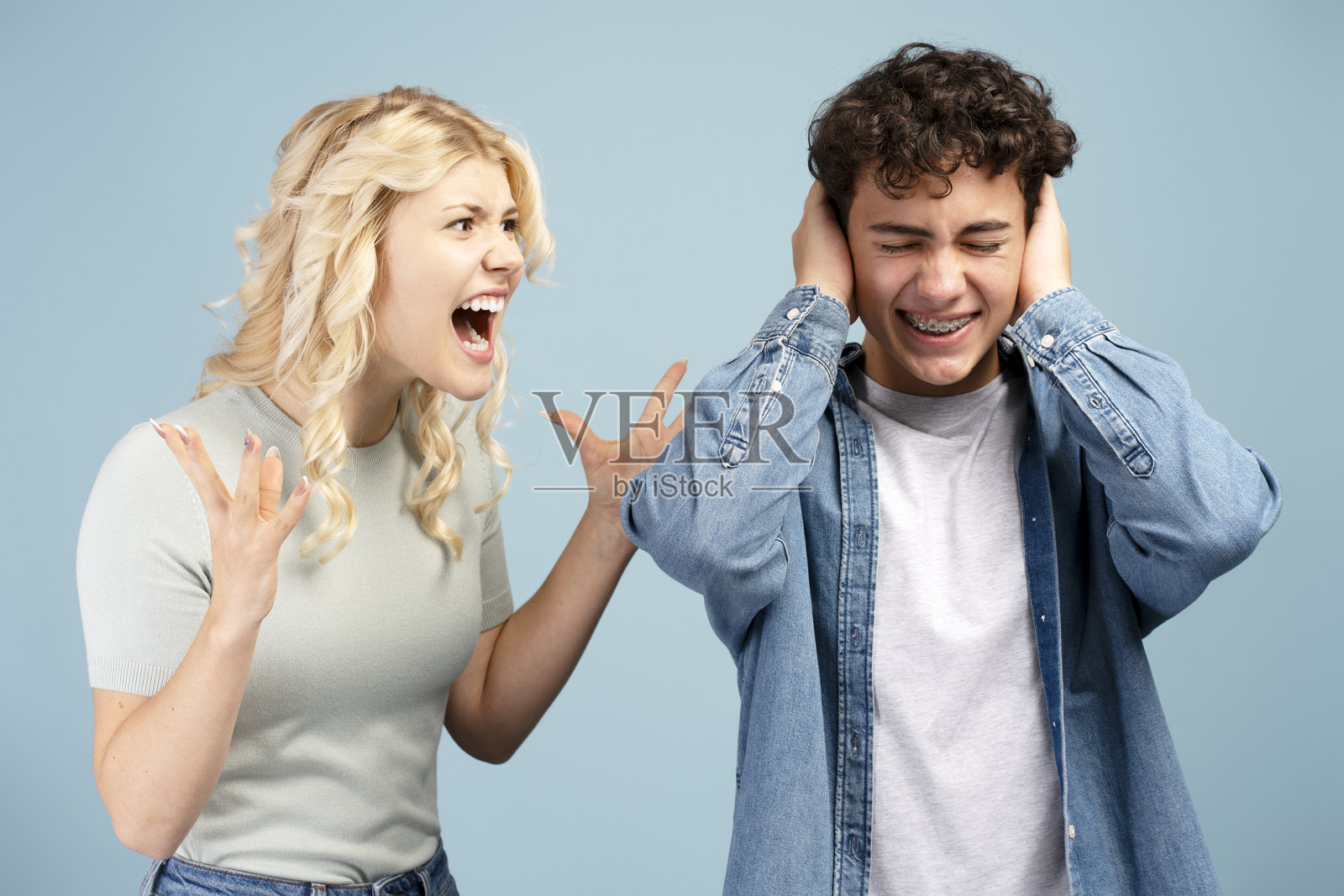 愤怒的小女孩对着男孩大喊，男孩捂着耳朵孤立地站在蓝色背景前。照片摄影图片