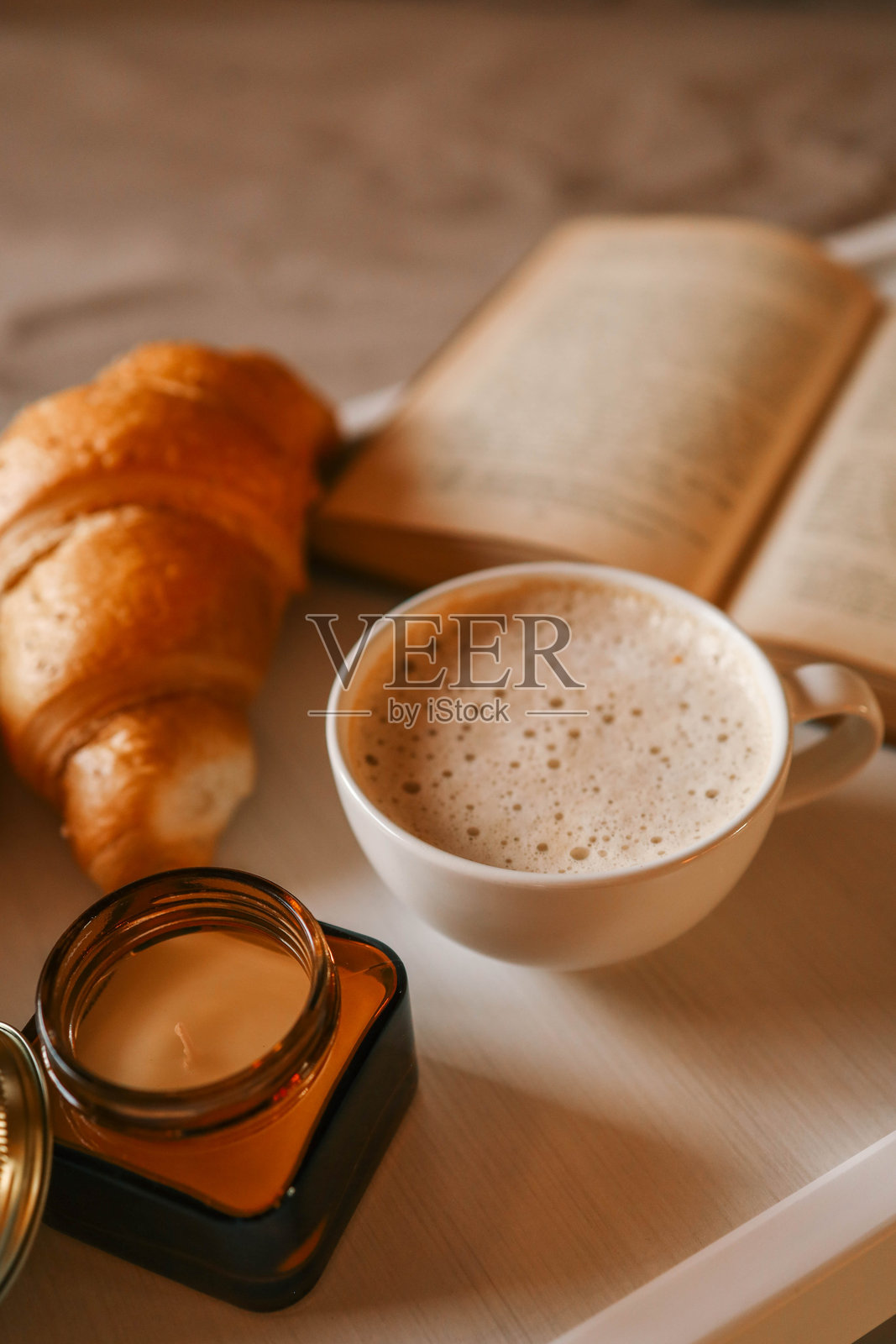 温馨的早餐场景特写，展示了一杯泡沫丰富的咖啡、金黄色的可颂面包和一个琥珀色的罐子，摆放在白色托盘上。柔和的阳光透过窗帘洒入，营造出温暖宜人的氛围。照片摄影图片