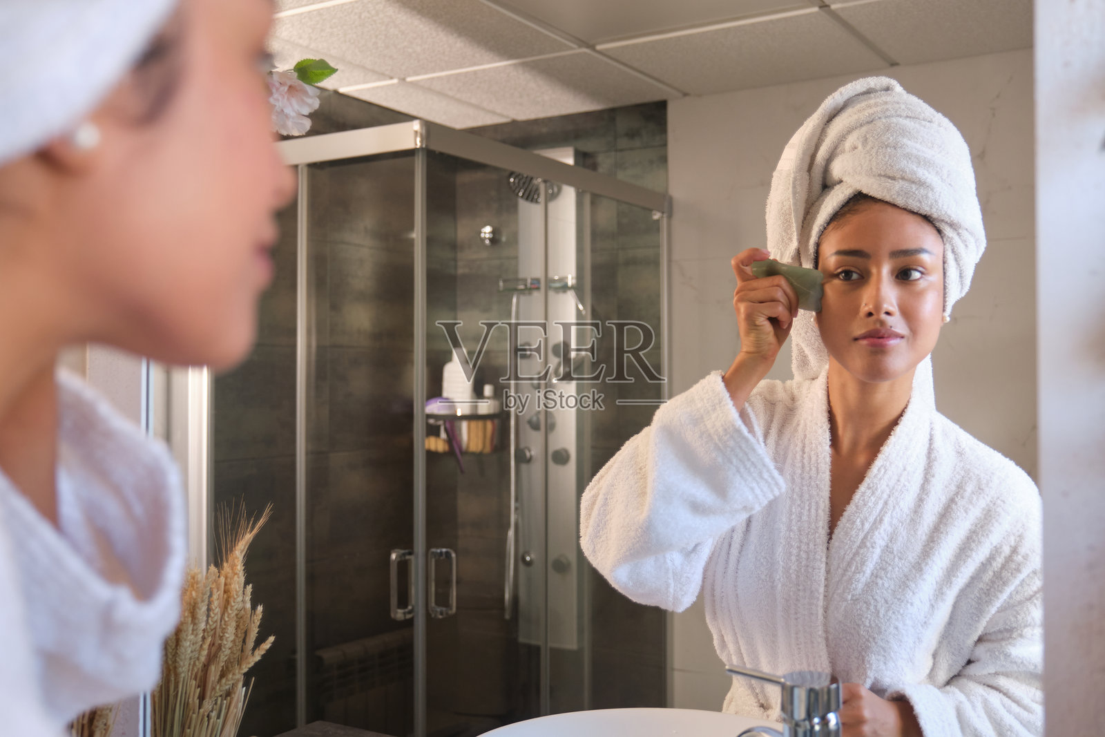 年轻女性在浴室镜子前使用刮痧石进行面部按摩照片摄影图片