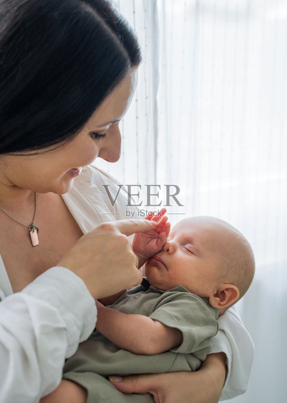妈妈站在窗边，怀抱着新生儿，轻轻触摸着熟睡儿子的手。关爱与支持，安睡与保护，母子之间的爱与关系。照片摄影图片
