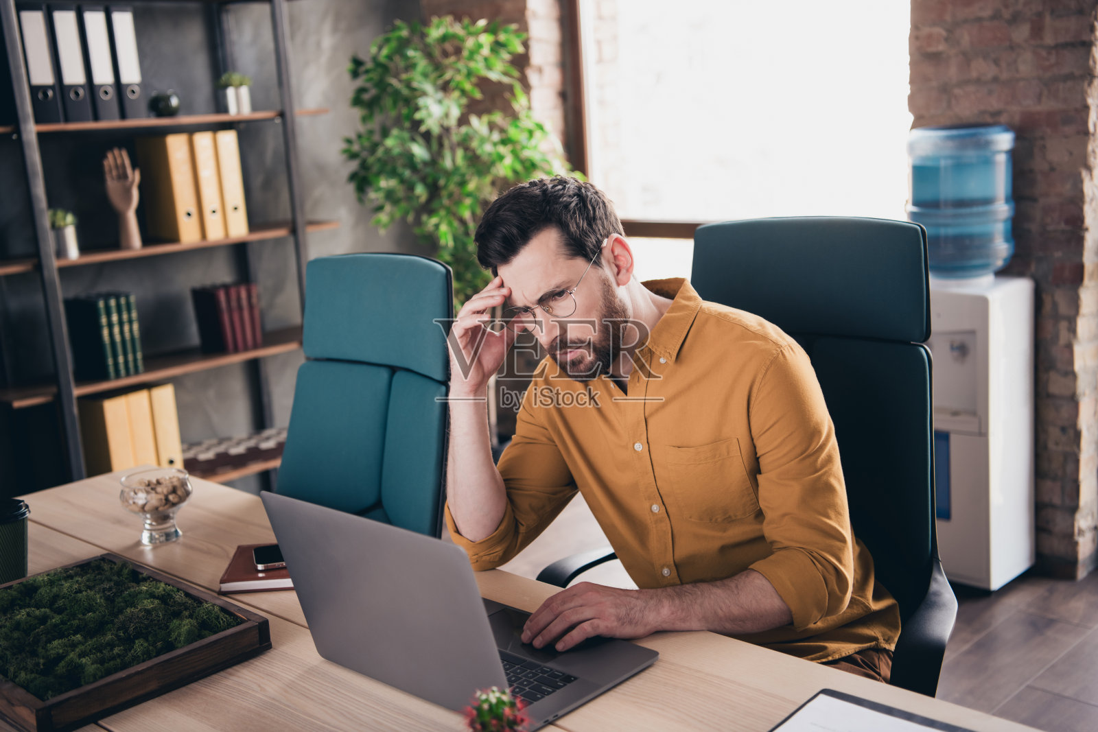 穿着黄色衬衫的年轻帅气男士在现代办公室阁楼内的工作空间里感到疲惫，正坐在笔记本电脑前。照片摄影图片