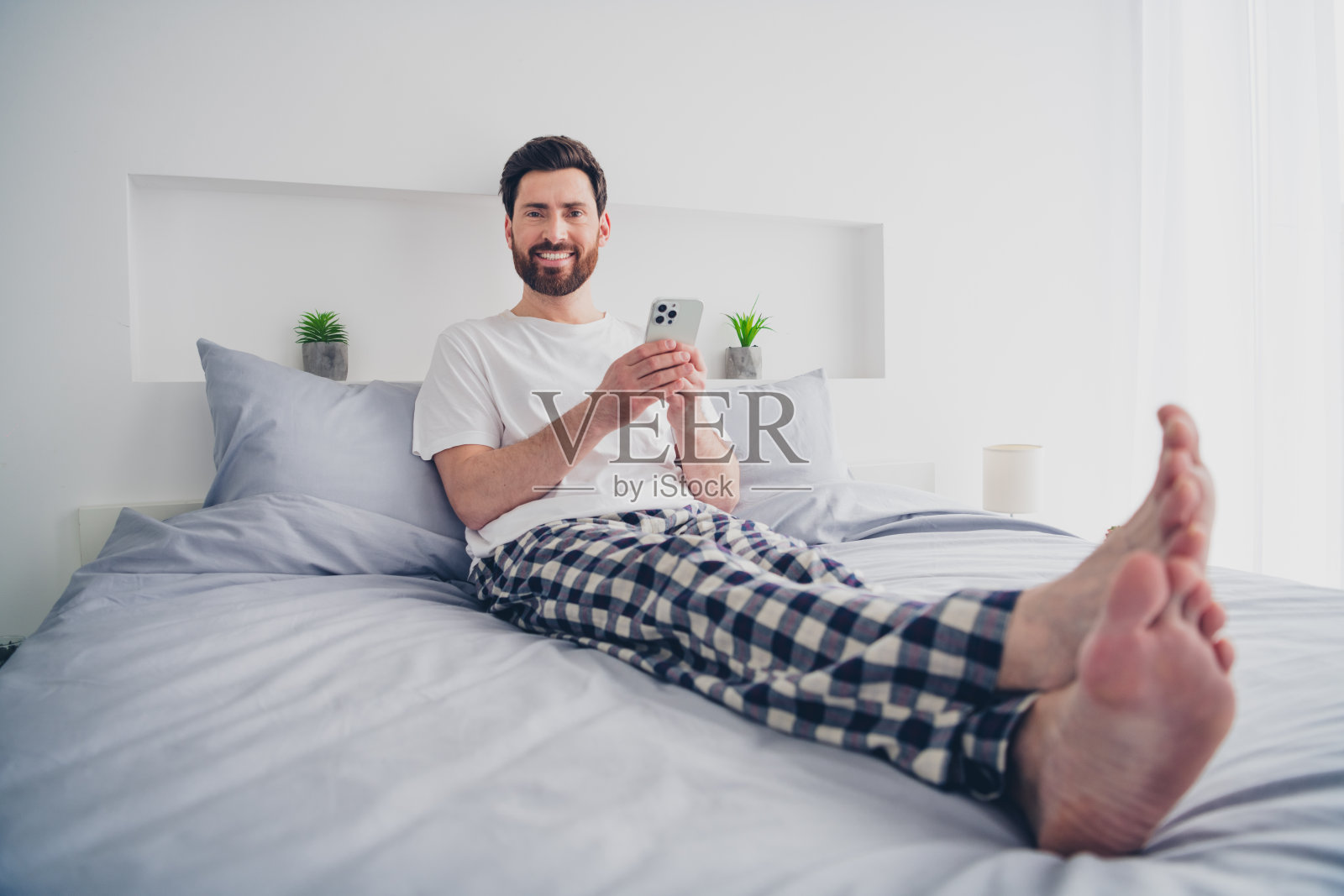 穿着睡衣的帅气成熟男士在室内床上躺着，心情愉快地用现代设备发短信。照片摄影图片