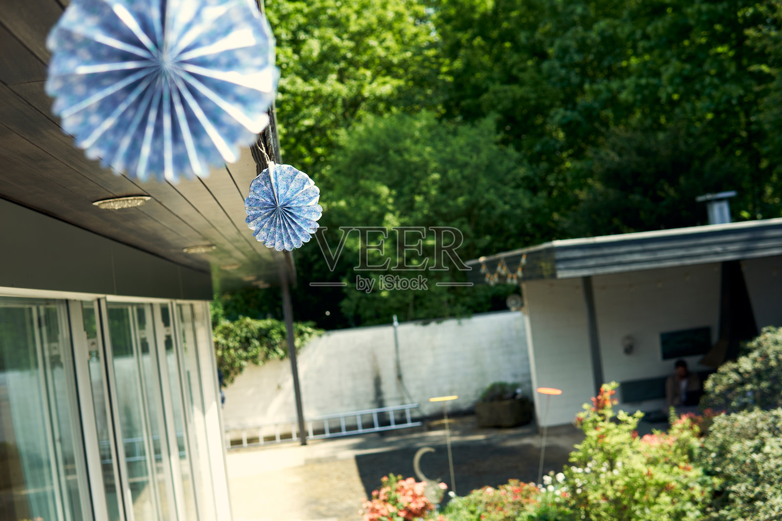 在阳光明媚的白天，装饰性纸扇悬挂在郁郁葱葱的花园环境中的建筑外。照片摄影图片