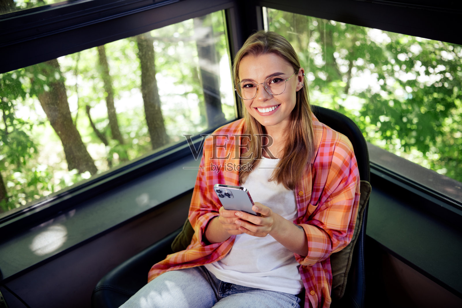可爱的年轻金发女士坐在桌子旁，手持电话，在咖啡馆、共享办公空间或图书馆工作，室内有大窗户，窗外可以看到公园的树木。照片摄影图片