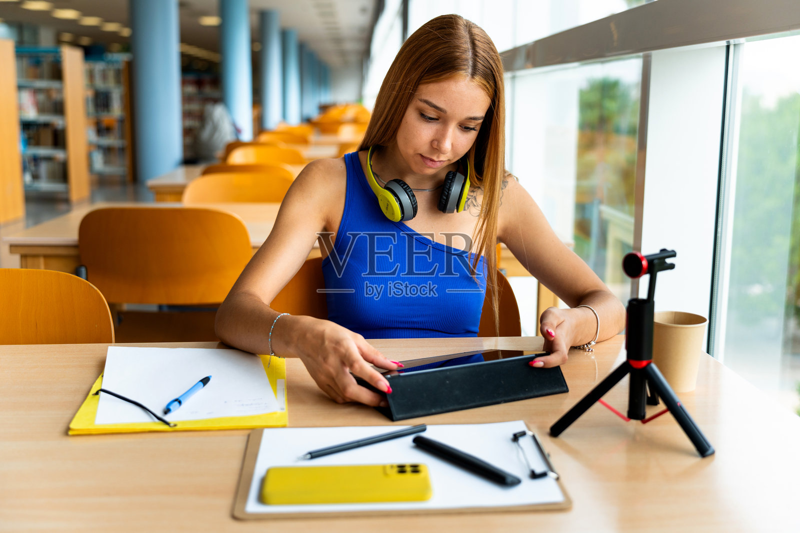 漂亮的大学女孩在图书馆学习。她在做笔记，使用耳机、平板电脑和手机。照片摄影图片