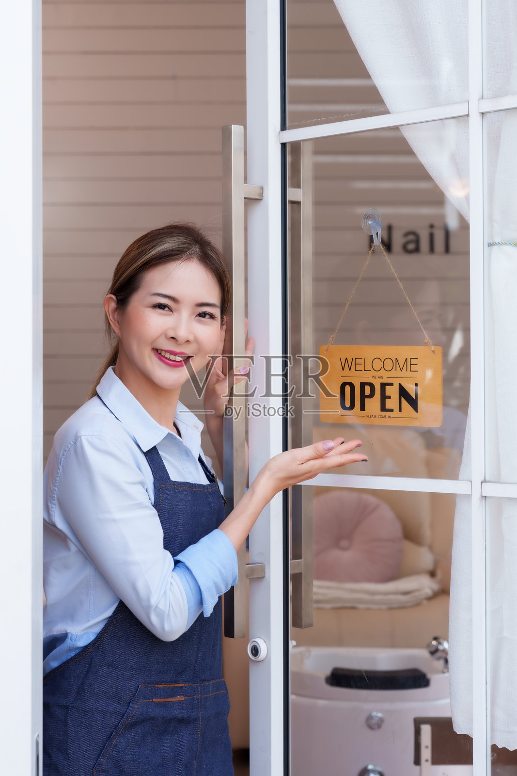 亚洲女性水疗中心老板在水疗中心入口处微笑，穿着蓝色围裙和衬衫，展示着开放标志。温馨、专业的水疗环境，准备迎接顾客享受健康和美容服务。照片摄影图片