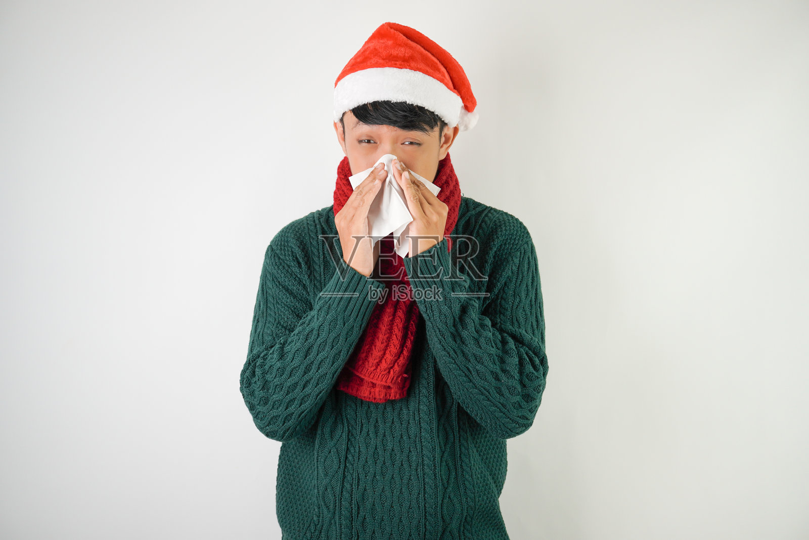 年轻的亚洲男子戴着圣诞老人帽子，系着红色围巾，穿着绿色长袖毛衣，正在打喷嚏并用纸巾遮掩，背景为白色。照片摄影图片