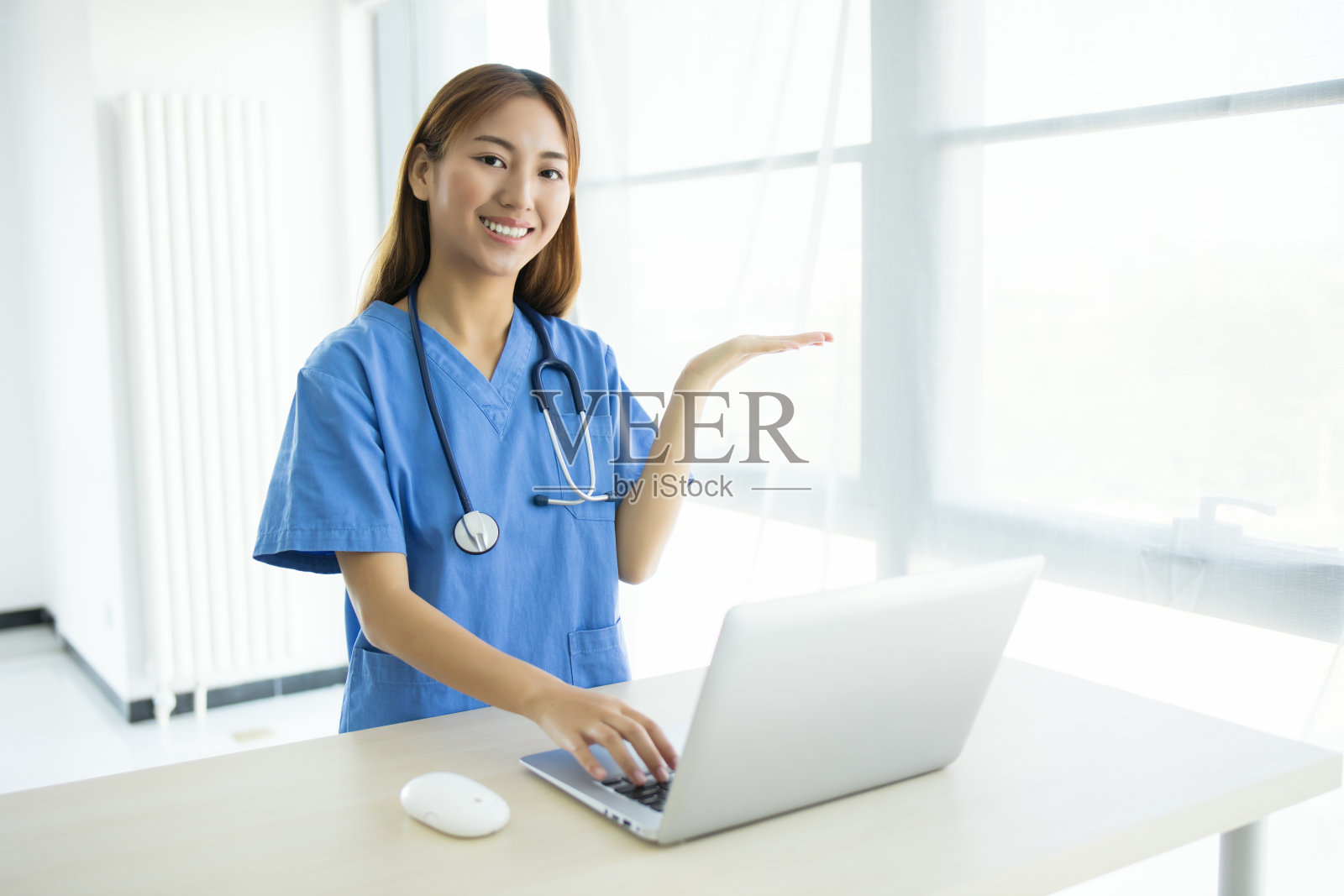 一位穿着蓝色制服的微笑女医生或护士坐在桌子旁使用笔记本电脑。照片摄影图片