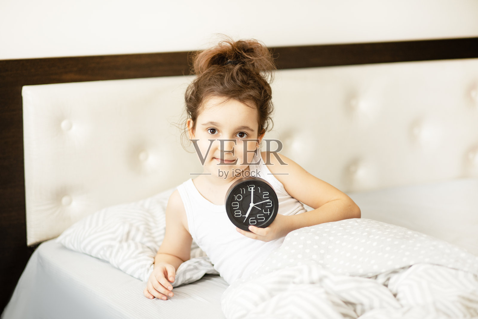 可爱的小女孩躺在床上，早晨醒来，手里拿着闹钟。照片摄影图片