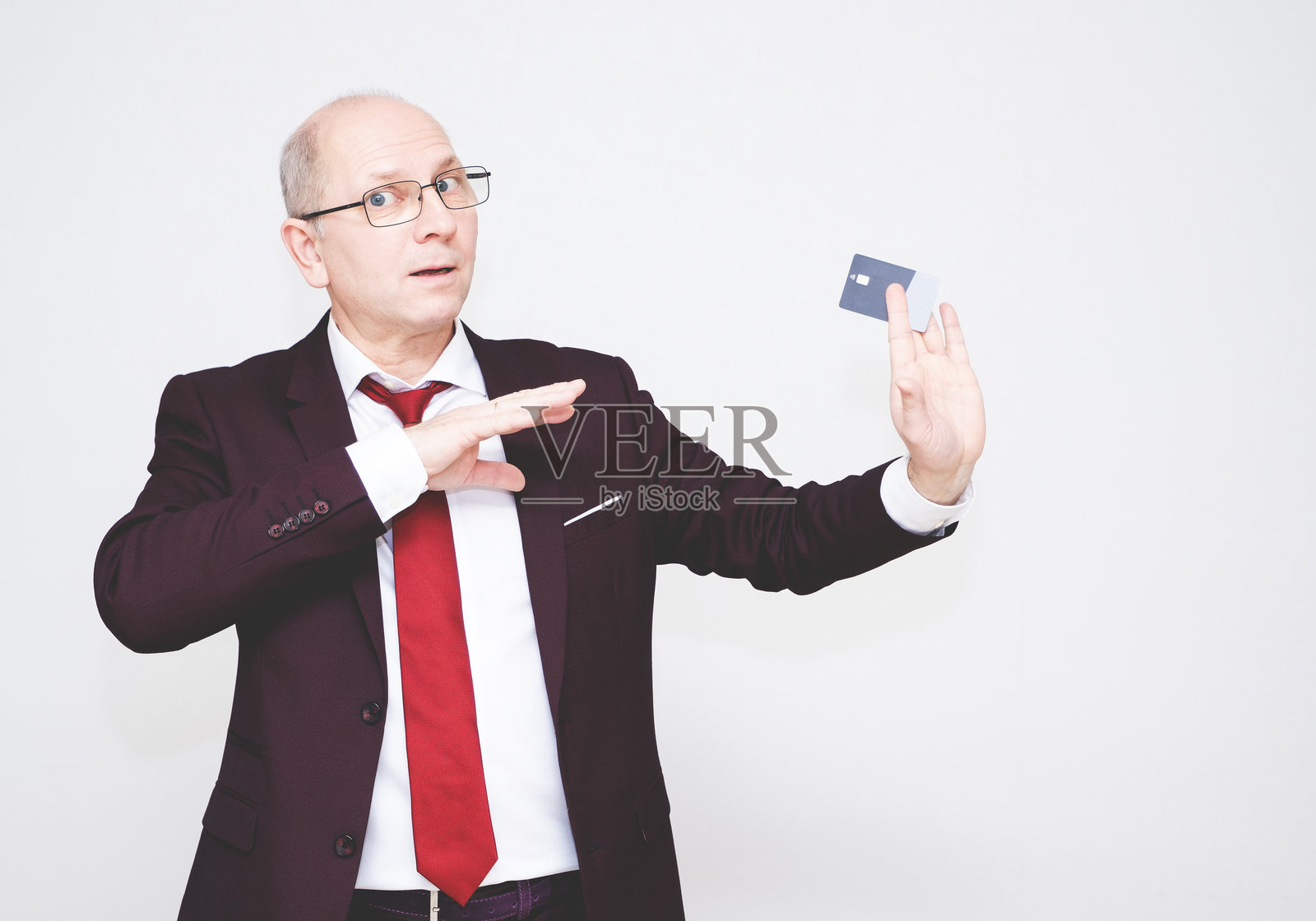 一位50岁的白人男性，穿着酒红色的商务西装，手中拿着一张信用卡，正在展示这张卡的把戏。照片摄影图片