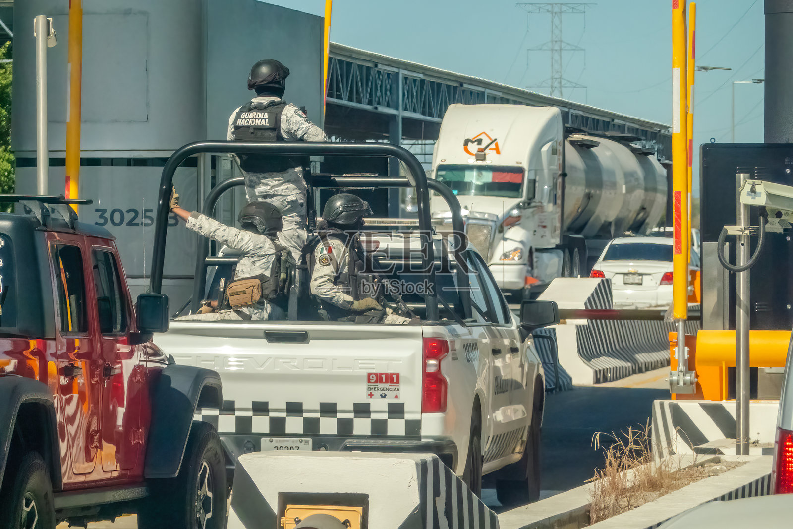 国民警卫队武装官员在一个检查站巡逻，驾驶着一辆白色皮卡车，旁边是其他卡车和车辆，天空晴朗。照片摄影图片