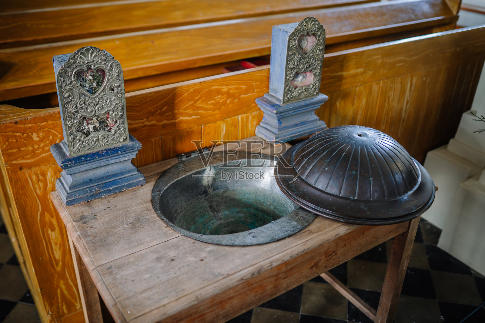 一个带有金属碗和盖子的洗礼盆，周围环绕着华丽的宗教装饰，放置在教堂的木长椅附近。照片摄影图片