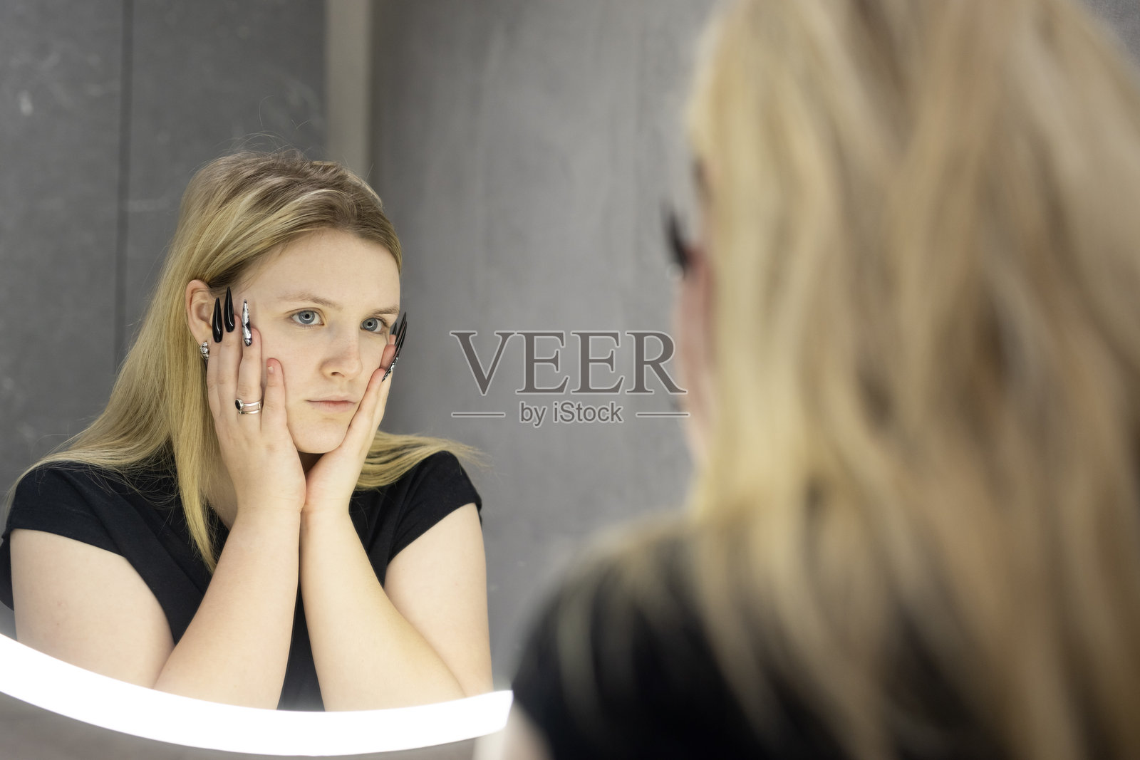 一位金发年轻女性在现代浴室的圆形照明镜前凝视自己的倒影，认真地调整自己的外貌。照片摄影图片