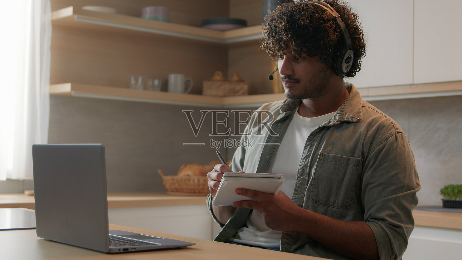 印度商人阿拉伯男教师戴着耳机，通过笔记本电脑进行视频通话会议，远程授课聊天，做笔记，男性在线导师在家厨房教学生学习互联网。照片摄影图片