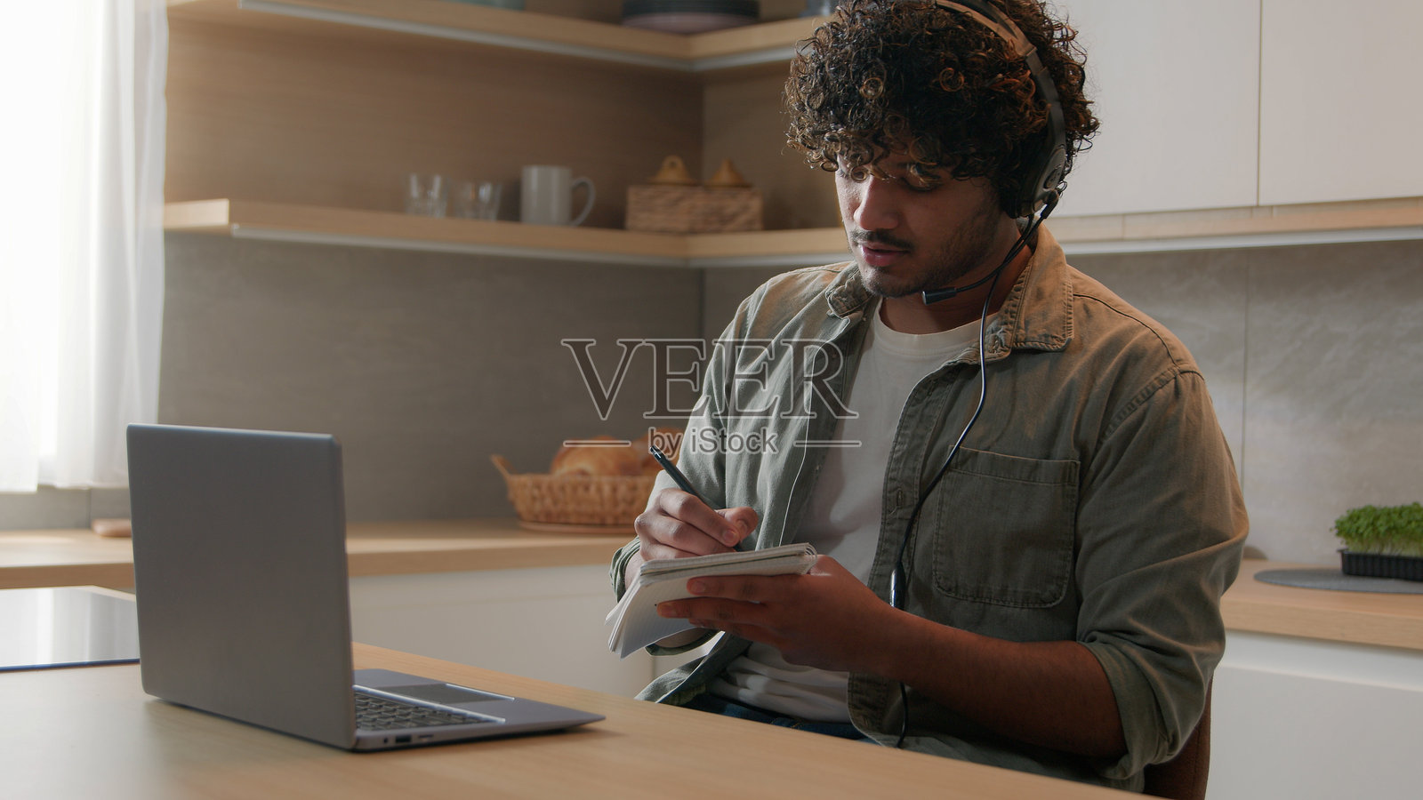 戴着耳机的西班牙裔男子在厨房里使用笔记本电脑参加远程会议，进行在线学习和辅导课程；阿拉伯男性商人在家中视频通话时写笔记。照片摄影图片