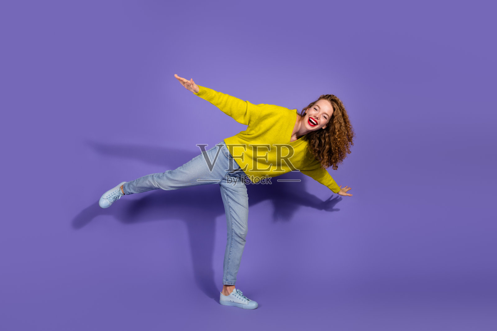 快乐的年轻女性在鲜艳的紫色背景前摆姿势，穿着黄色毛衣和牛仔裤，展现出快乐与时尚。照片摄影图片