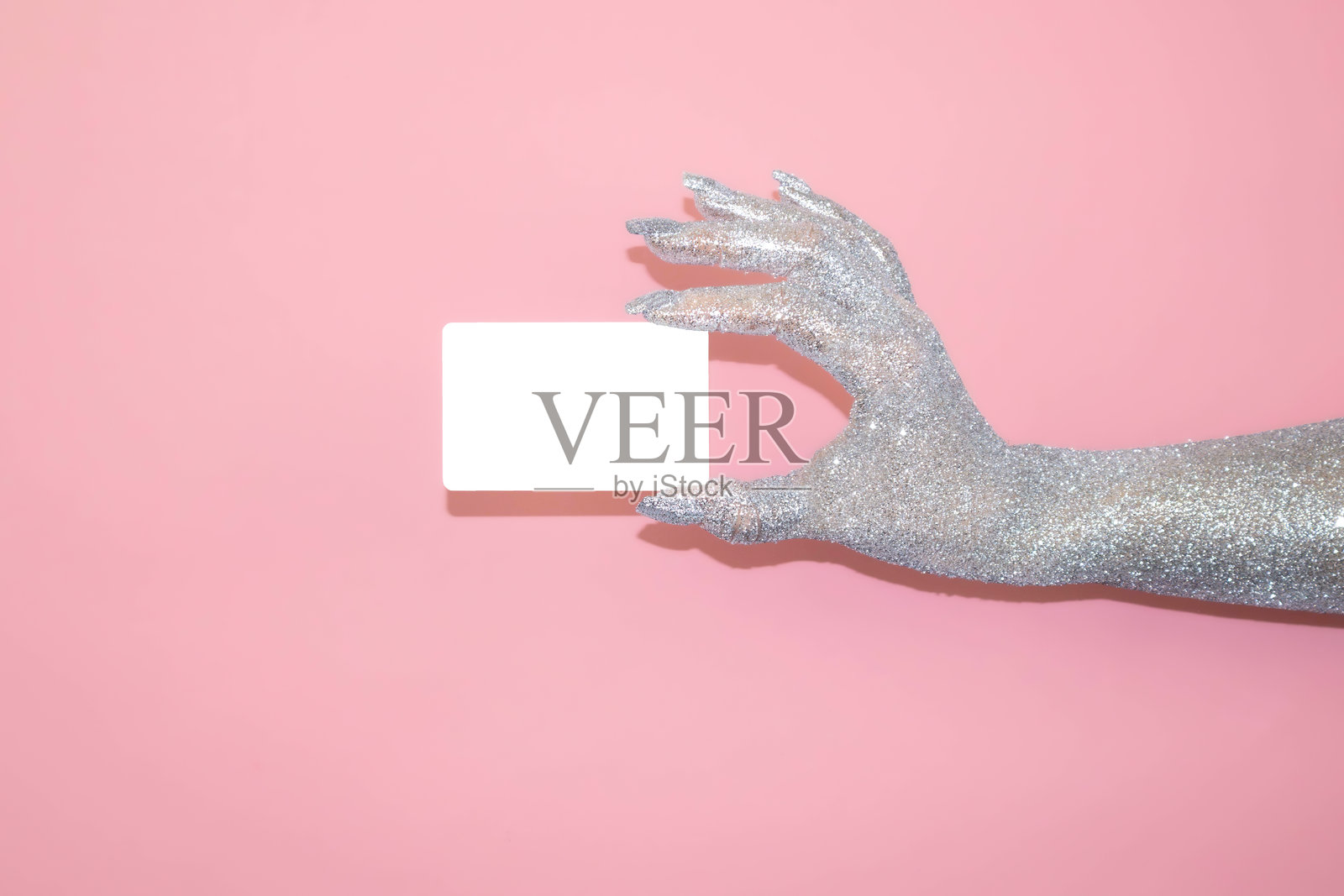 闪闪发光的银色手握着一张空白卡片，背景为粉色。照片摄影图片