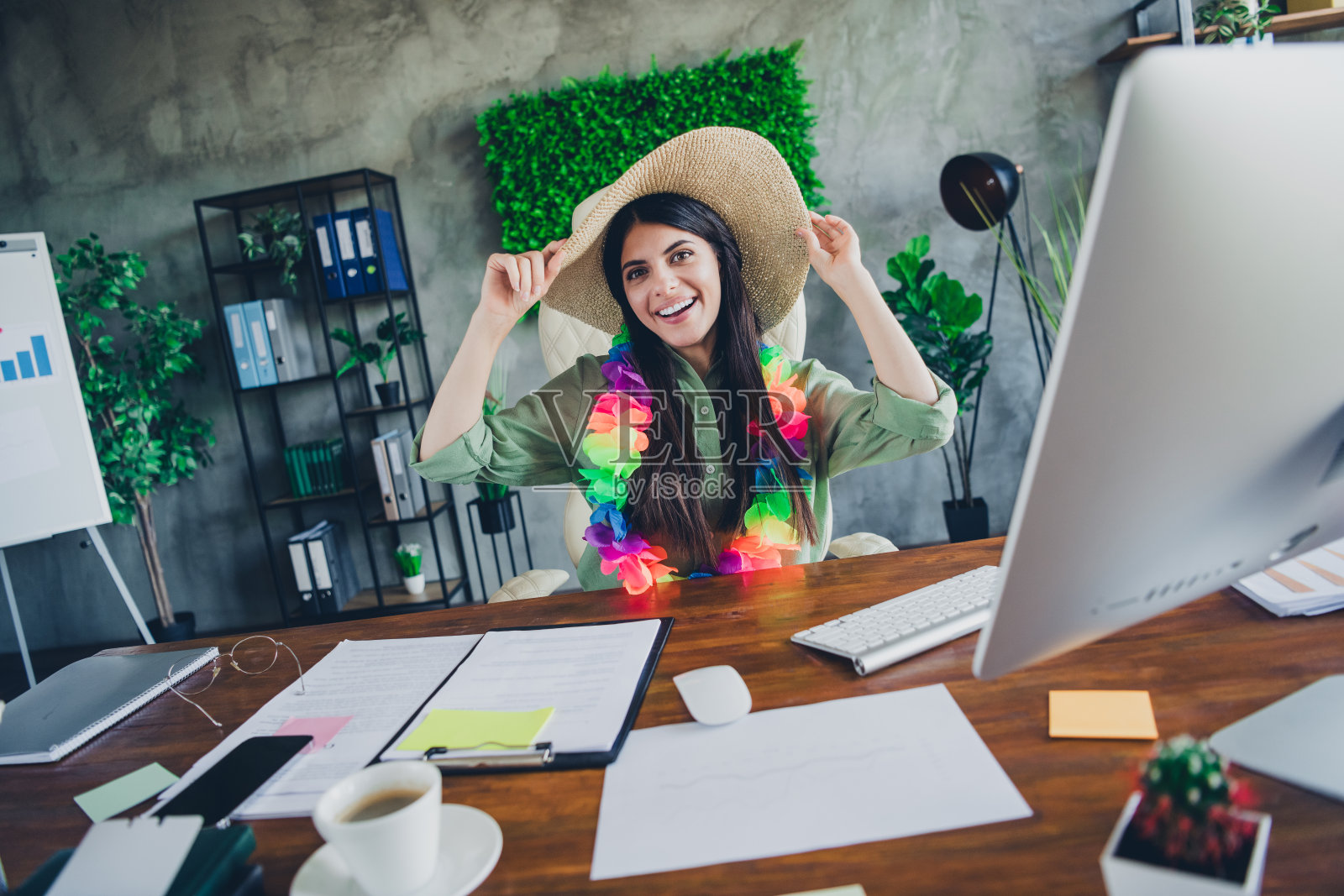 快乐的女商人留着棕色头发，戴着夏季帽子和五彩缤纷的花环，在现代办公室中微笑，周围有生机勃勃的绿色植物。照片摄影图片