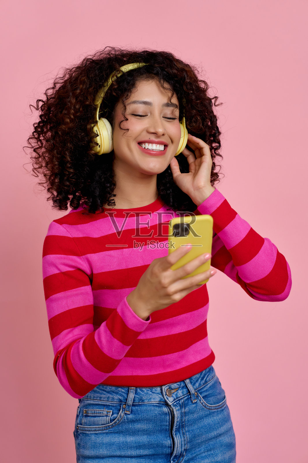 快乐的拉丁裔女性在粉色背景下戴着黄色耳机，使用智能手机听音乐照片摄影图片