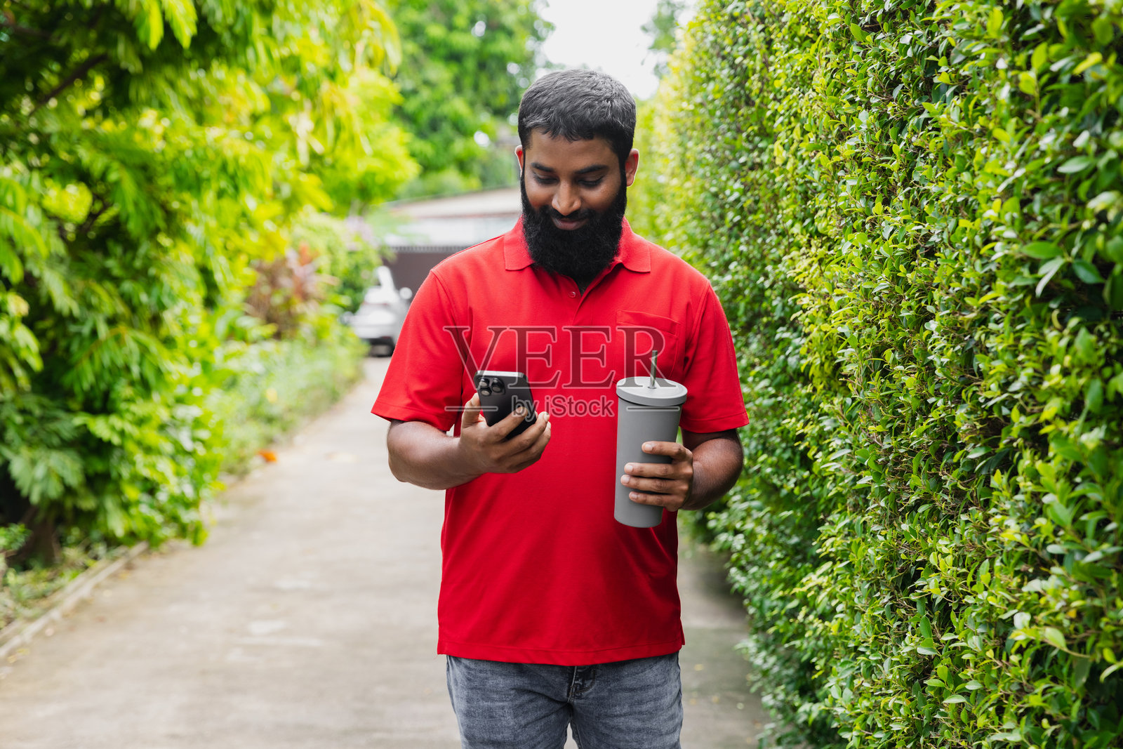 一个留着胡子的男人在阳光明媚的日子里走在郁郁葱葱的小路上查看手机。照片摄影图片