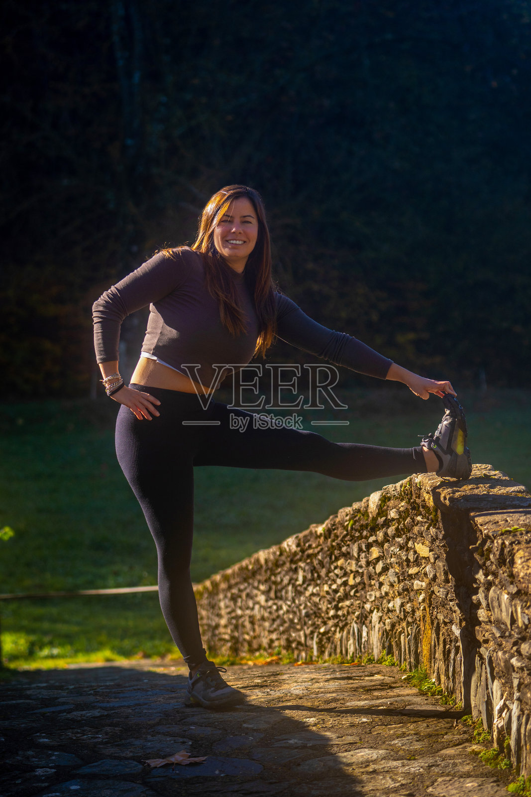 年轻女性穿着紧身运动服，在乡村桥的石墙上伸展腿部，微笑着看向镜头，进行户外训练，背景是日出时的山脉。照片摄影图片