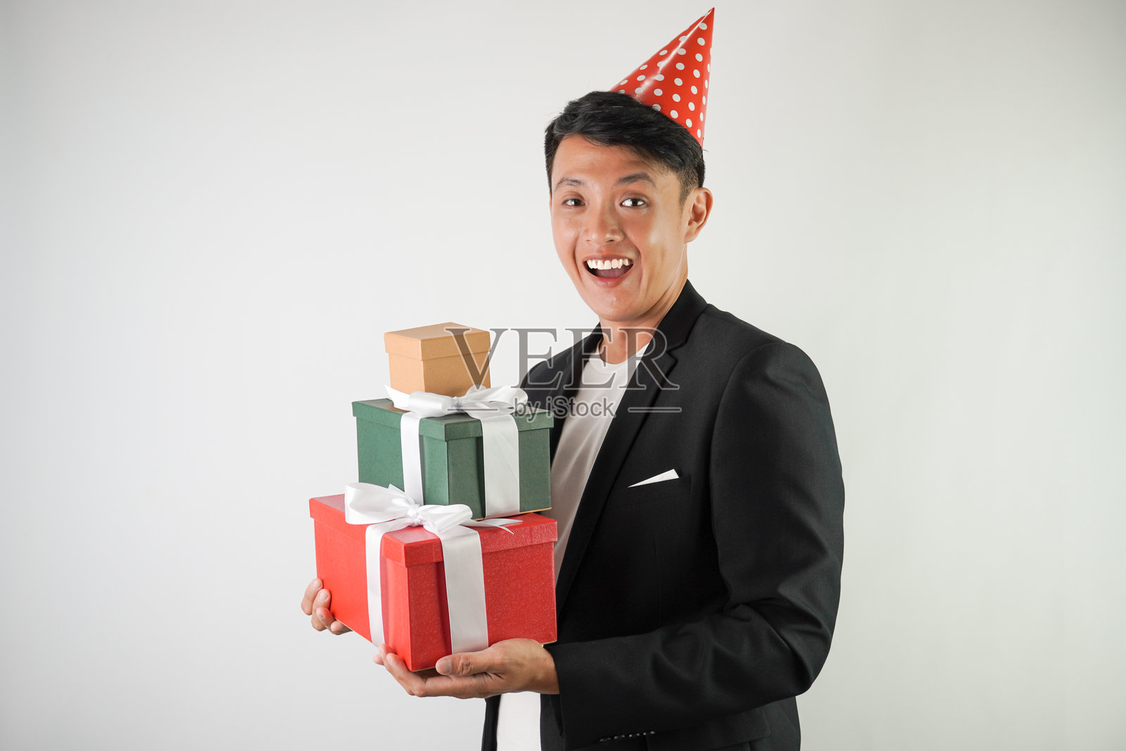 兴奋的年轻亚洲男子戴着红色派对帽，穿着白衬衫和黑色西装，手里提着一个带丝带的大礼盒，背景为白色。适用于圣诞节和新年派对的概念。照片摄影图片