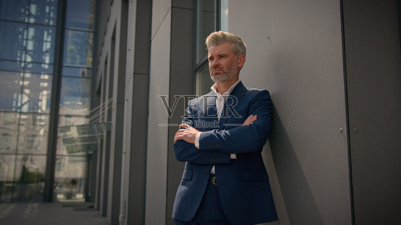 户外肖像，沉思的白人老年男性，成熟的商业经理，中年雇主，年长的商人，微笑着看向镜头，双手交叉，靠在城市的墙上。照片摄影图片