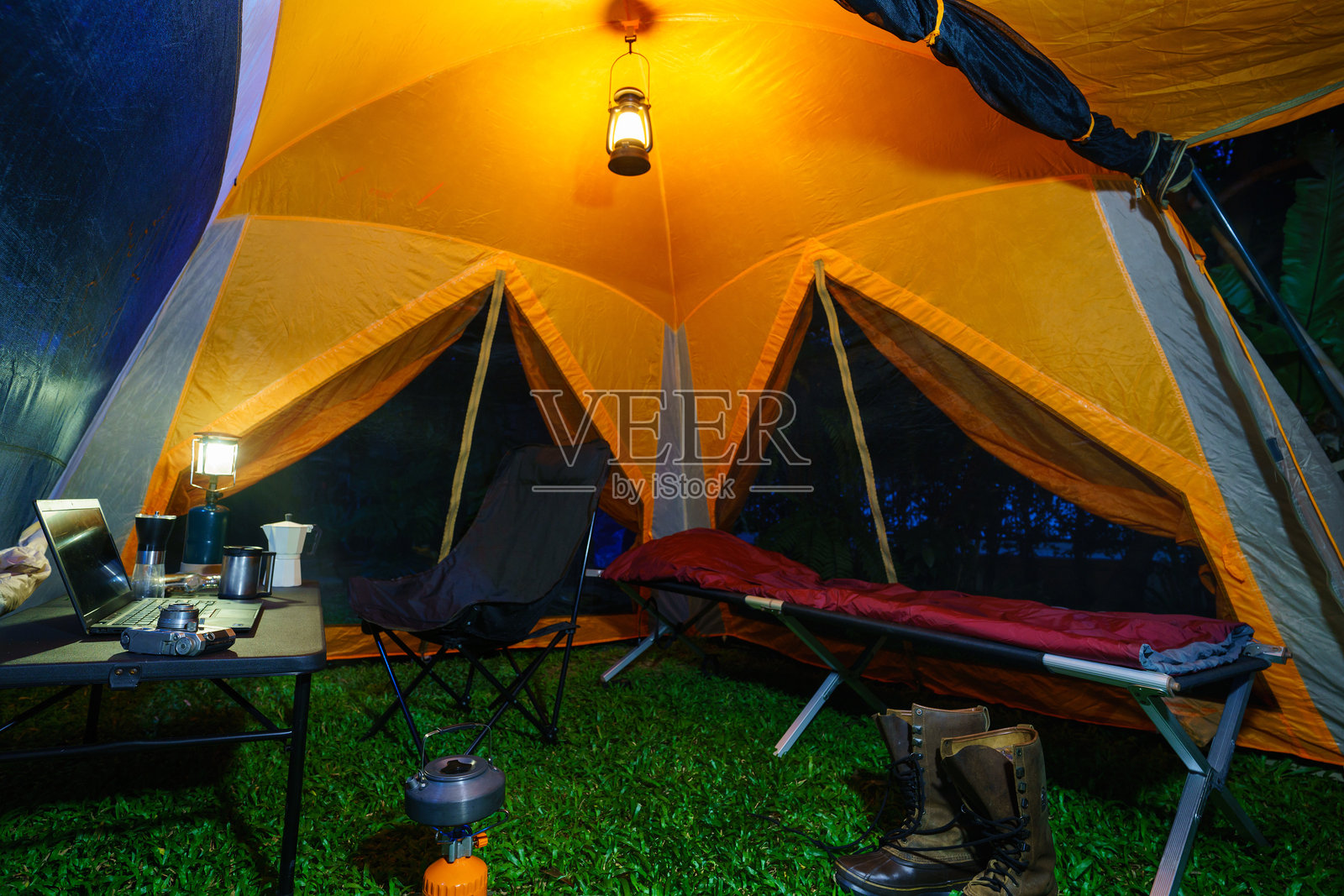 森林夜晚的露营帐篷内有折叠床、煤气灯和咖啡套装，木质露营桌上放着笔记本，以及户外创意。照片摄影图片