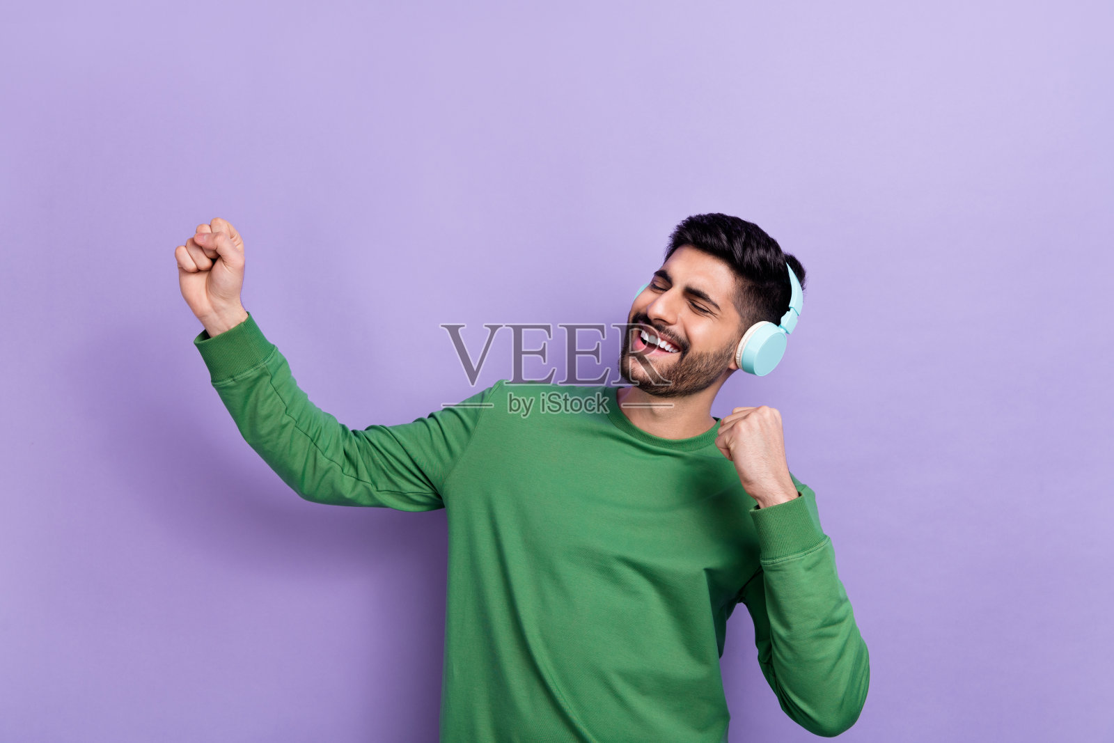 愉快的男孩穿着绿色长袖，戴着耳机，唱着最喜欢的艺术家的歌，双拳高举，背景为紫色。照片摄影图片