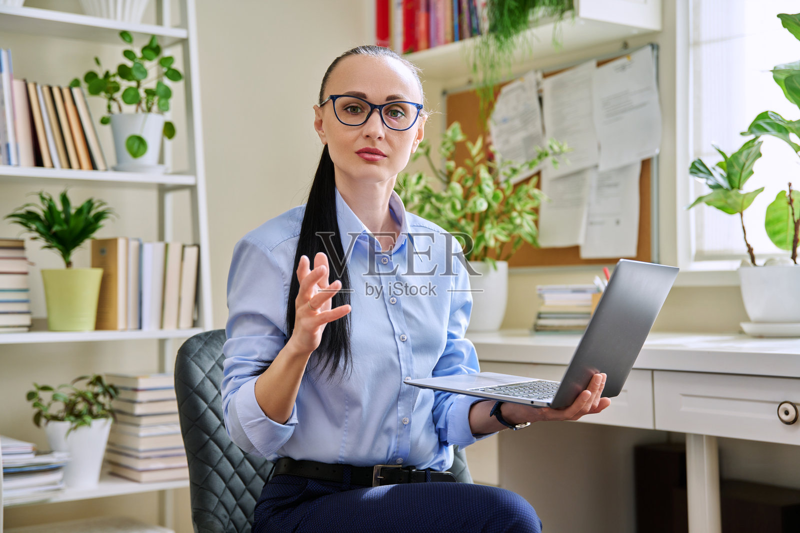 中年商务女性手持笔记本电脑，严肃地看着镜头，坐在家中办公室里照片摄影图片