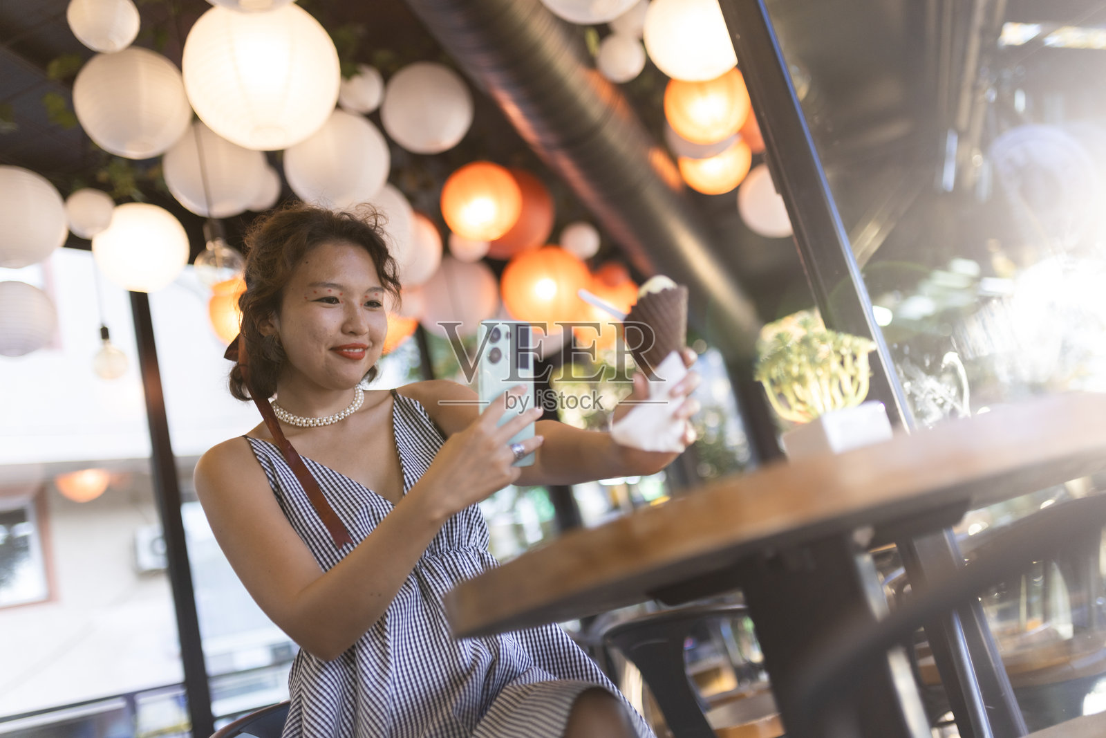 年轻女性在一家装饰着五彩灯笼、温馨宜人的时尚咖啡馆里享受冰淇淋时自拍。照片摄影图片