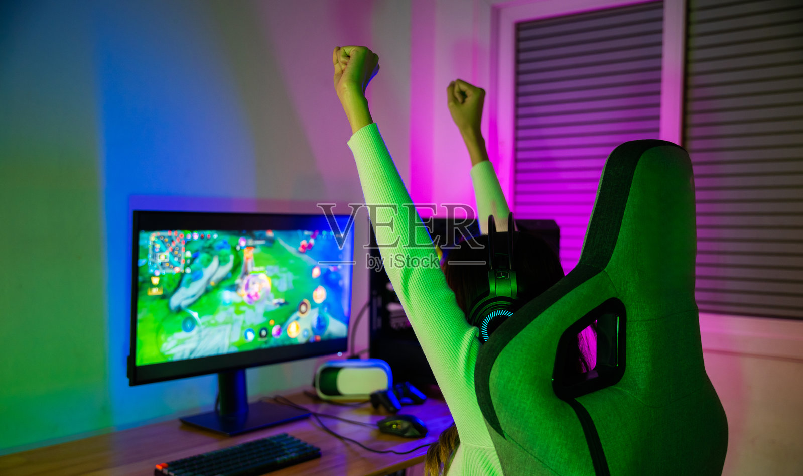 获胜。年轻女性戴着游戏耳机在家中霓虹灯房间里玩在线视频游戏，感到兴奋。快乐的玩家年轻人正在玩在线电脑游戏，她举起双手庆祝赢得比赛。照片摄影图片