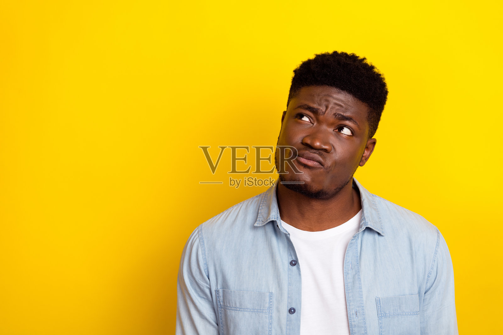 年轻的棕发男士思考着，穿着牛仔裤和衬衫，背景为黄色。照片摄影图片