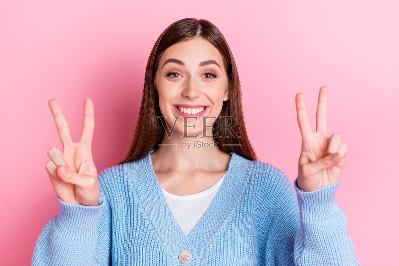 特写照片，迷人而无忧无虑的女孩穿着蓝色毛衣，做出V字手势，背景为粉色。照片摄影图片