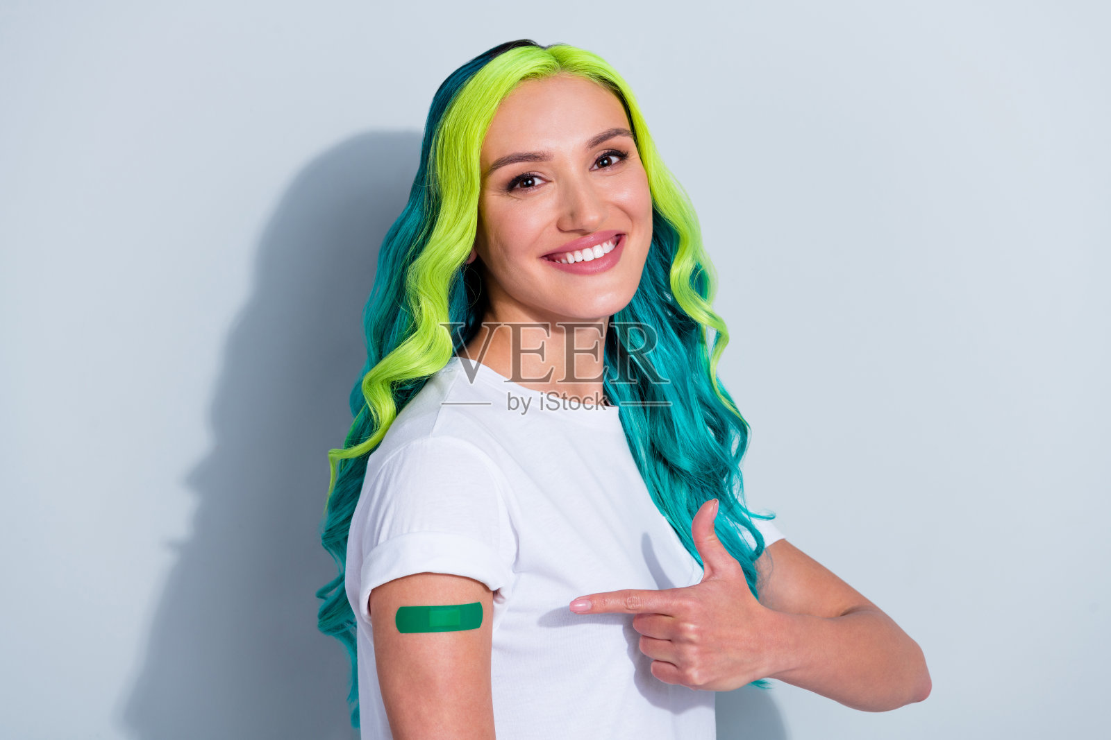 穿着T恤的可爱女孩，拥有甜美的渐变色头发，指着新冠病毒创可贴，背景为白色。照片摄影图片