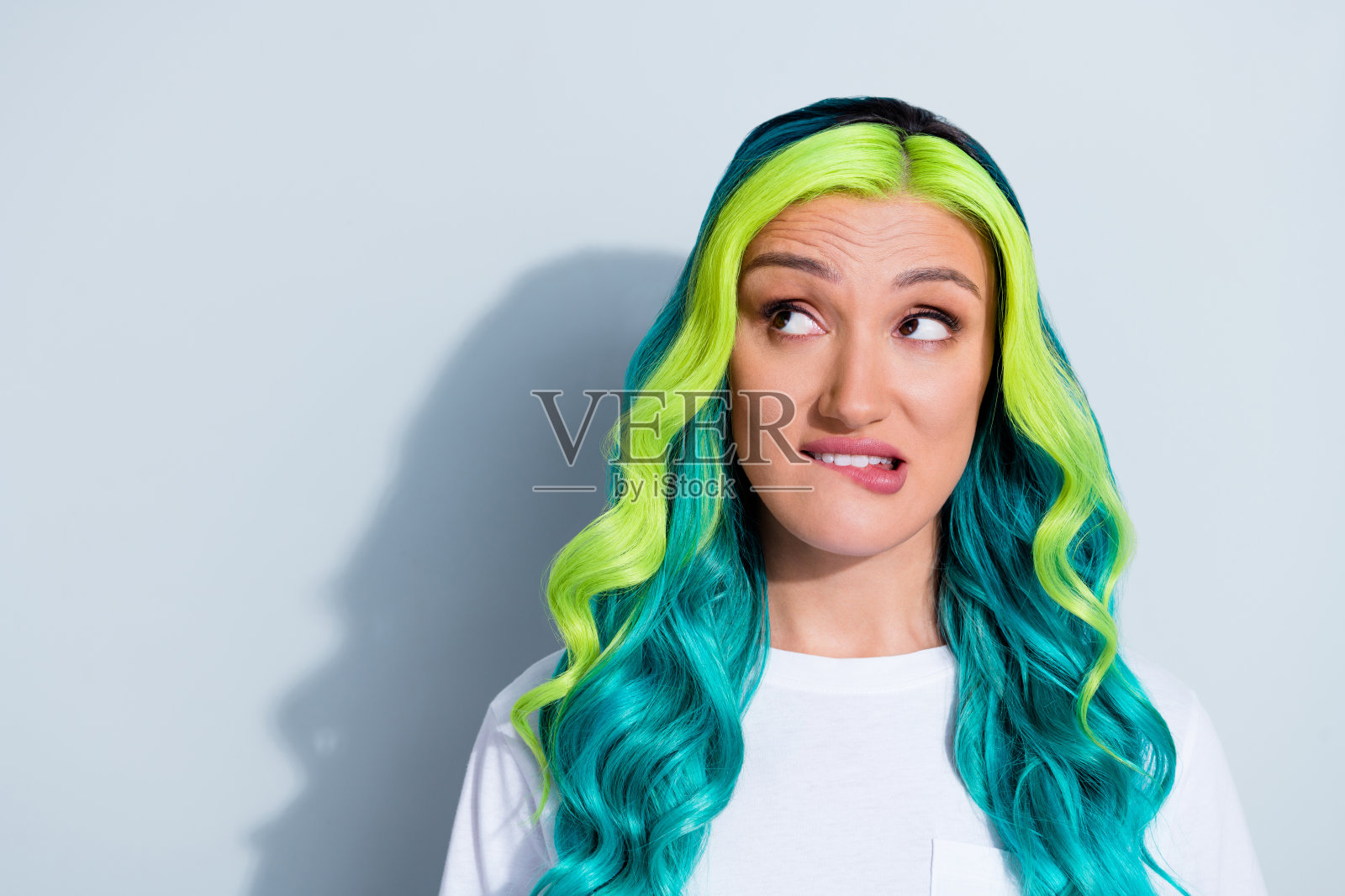 可爱的、不确定的女孩，拥有鲜艳渐变的头发，穿着T恤，咬着嘴唇，目光空洞，背景为白色。照片摄影图片