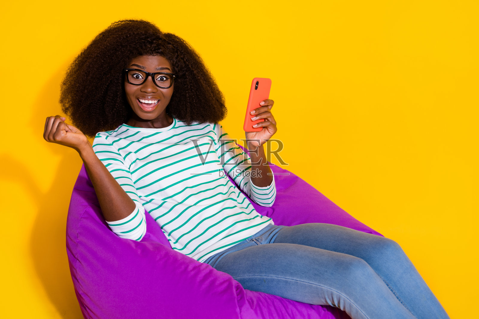 高兴的女孩坐在包上，举起拳头庆祝好运，手持电话，背景为黄色。照片摄影图片