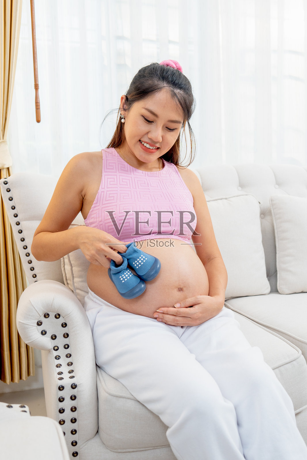垂直图像显示一位亚洲孕妇坐在沙发上，手中拿着一双蓝色鞋子，轻轻触碰右侧的肚子，脸上洋溢着幸福的微笑。照片摄影图片