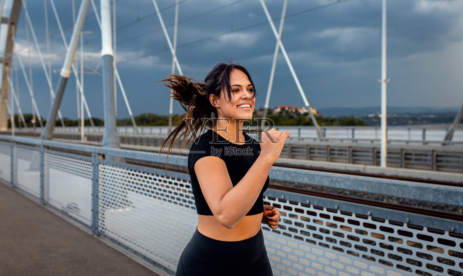 年轻女性穿着黑色运动服在日落时分的城市桥上跑步。照片摄影图片