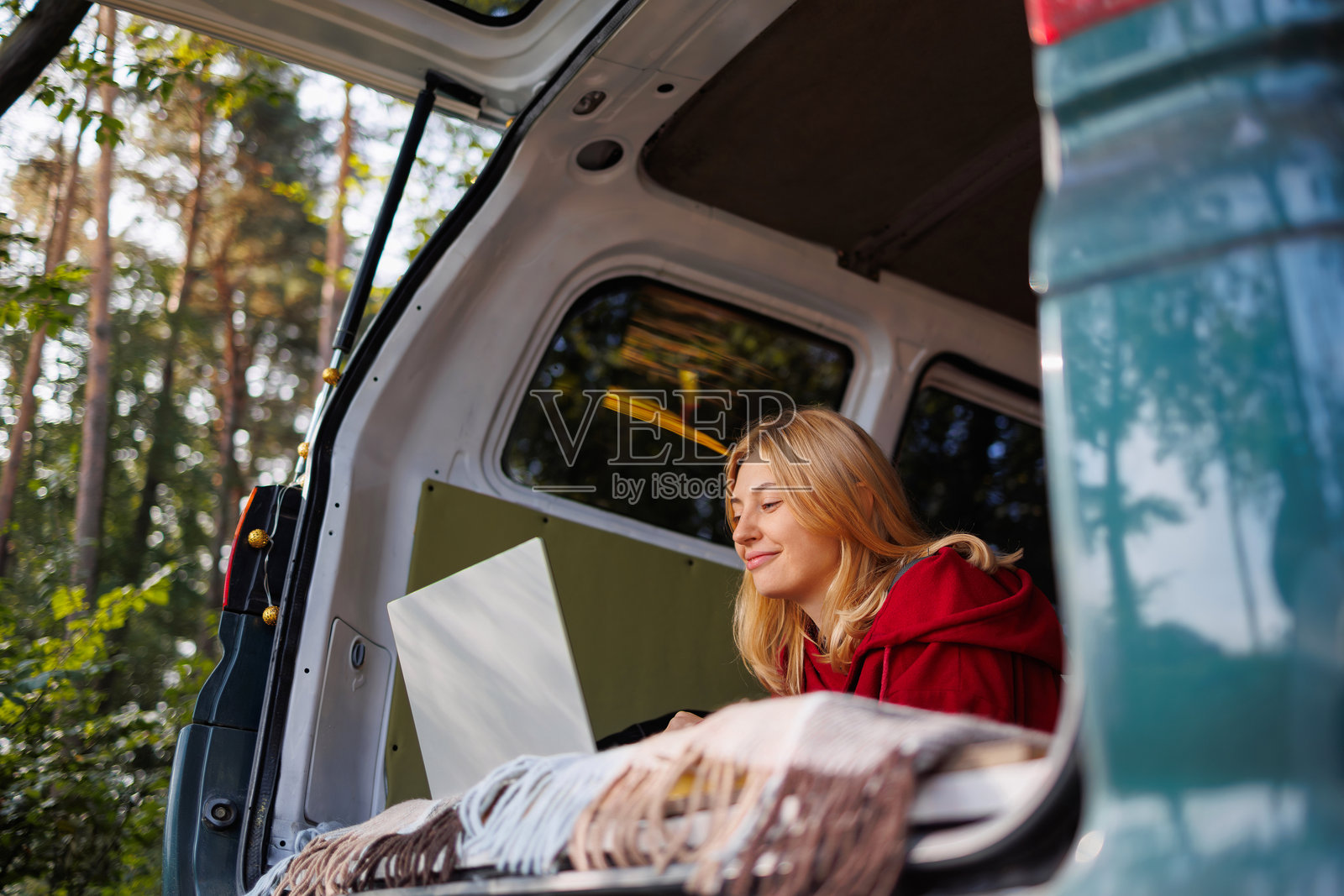 红发年轻女性穿着红色连帽衫，躺在露营车里使用笔记本电脑，周围是大自然。照片摄影图片