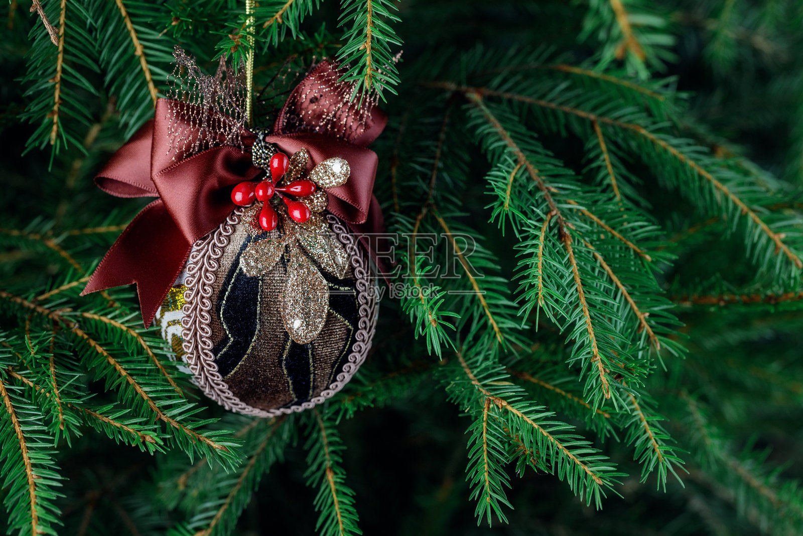 一个用棕色天鹅绒制成的圆形圣诞树玩具，配有酒红色丝带，装饰着金色细节和红色浆果，悬挂在圣诞树的树枝上。照片摄影图片
