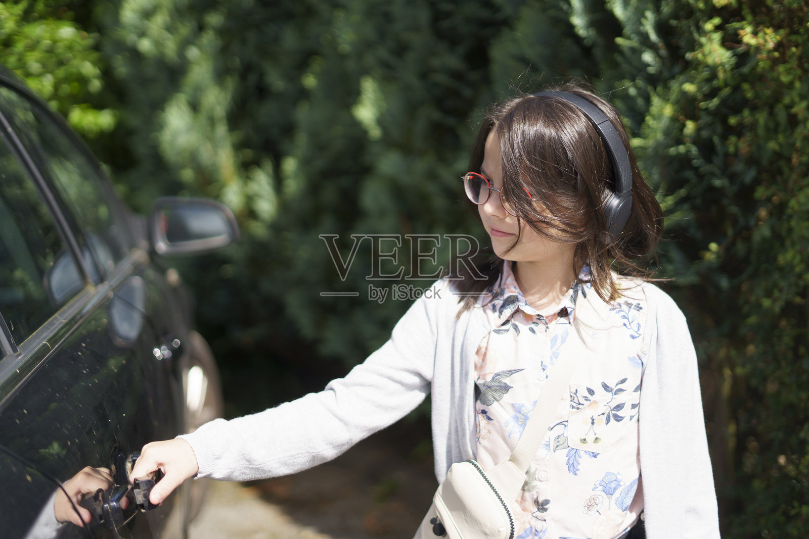 戴着大耳机和眼镜的小女孩站在夏天的车旁，手握车门把手。儿童旅行、活动、乐趣和快乐的概念。照片摄影图片