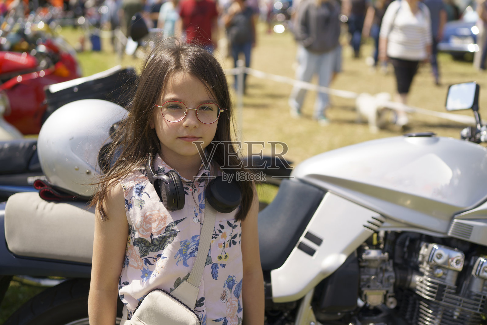 戴着眼镜和耳机的小女孩在老式汽车和摩托车的背景下玩得很开心。儿童旅行、活动、乐趣和获得新体验的概念。照片摄影图片