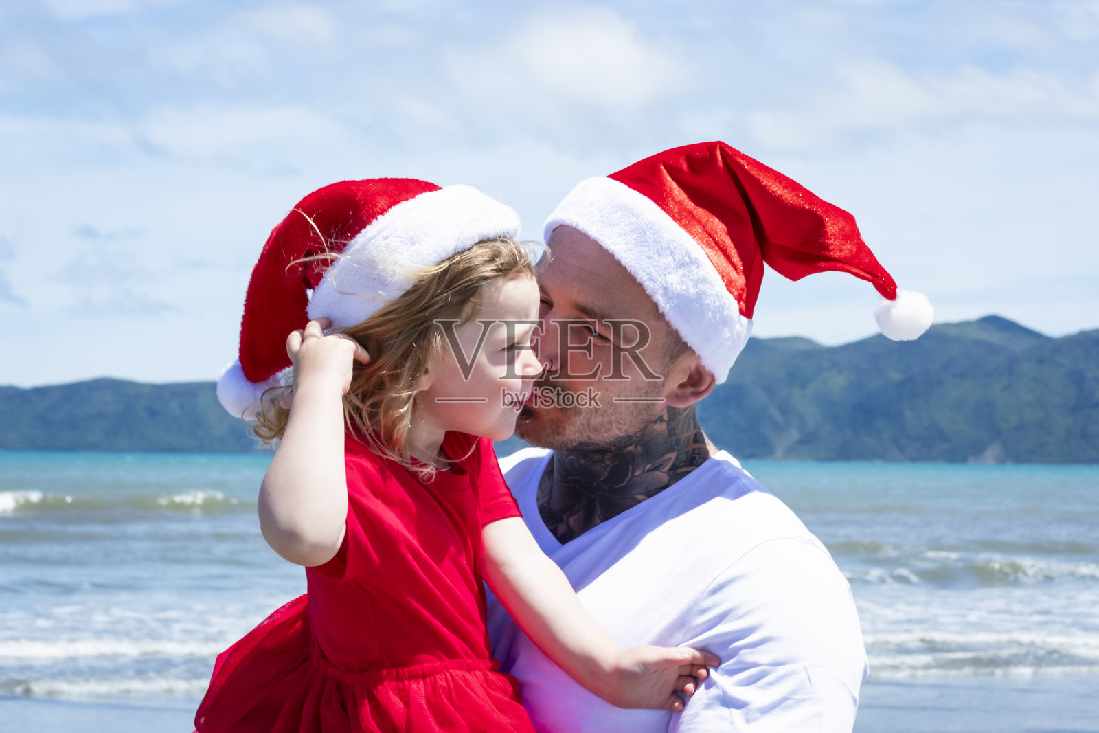 一个年轻女孩和她的父亲在海滩上度过美好时光，戴着圣诞帽。照片摄影图片