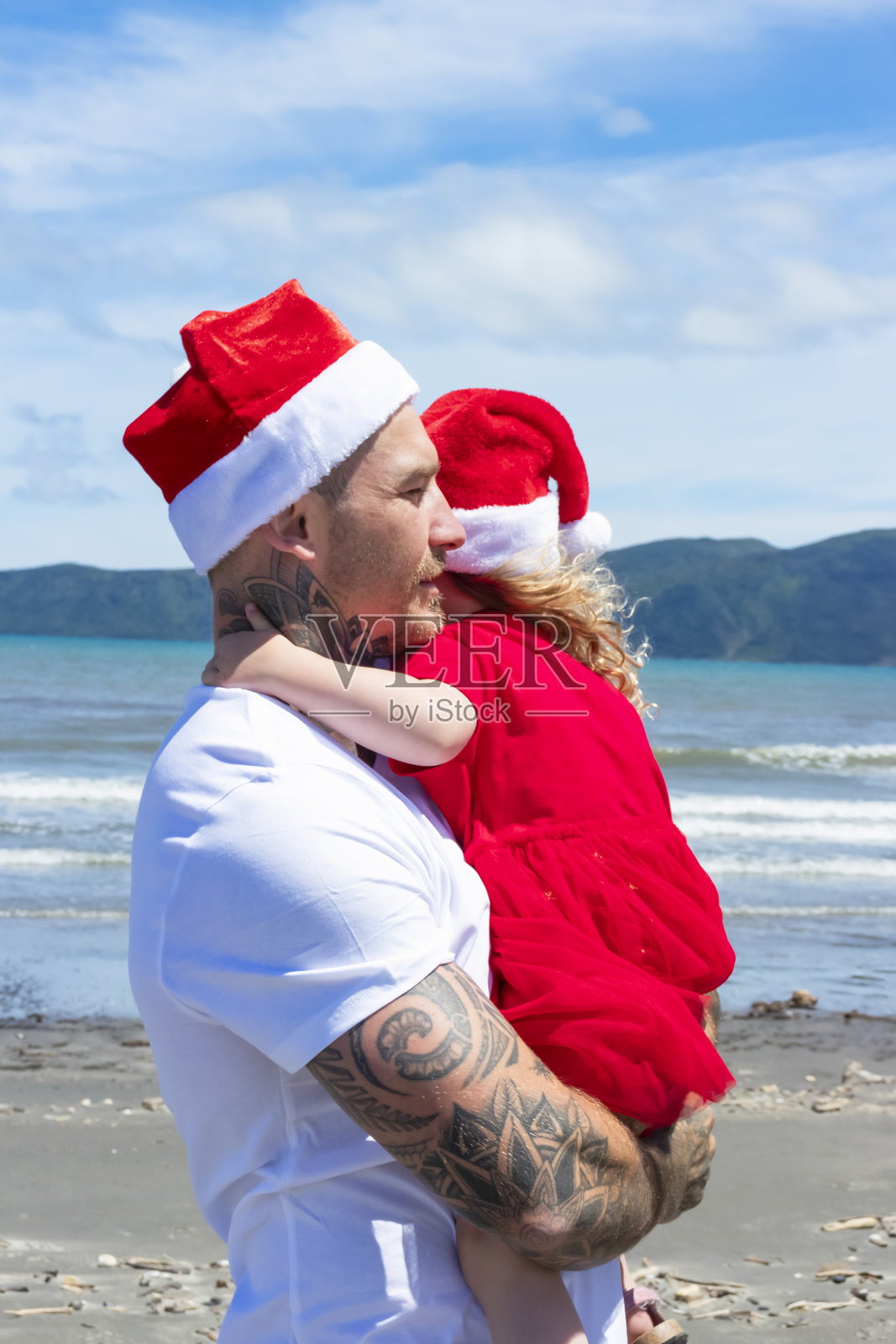 一个年轻女孩和她的父亲在海滩上度过美好时光，戴着圣诞帽。照片摄影图片