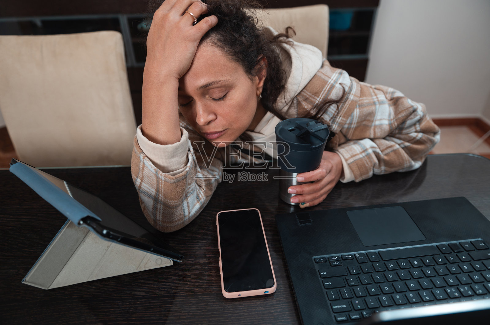 疲惫的女性坐在桌前，面前有笔记本电脑、手机和咖啡杯照片摄影图片