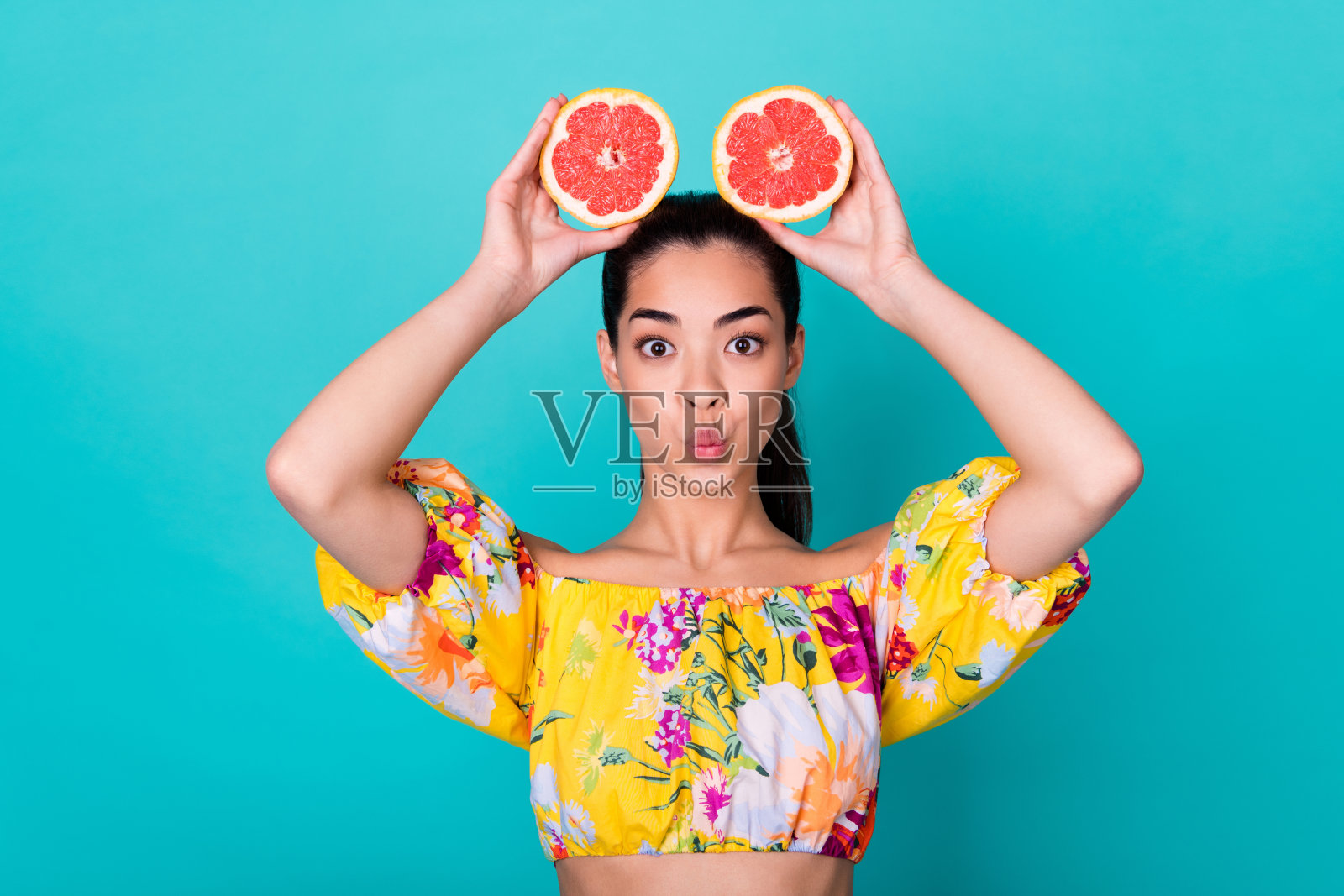 年轻可爱的中国女孩做鬼脸，嘴唇撅起，手里拿着切开的葡萄柚，耳朵旁边，背景为青色。照片摄影图片