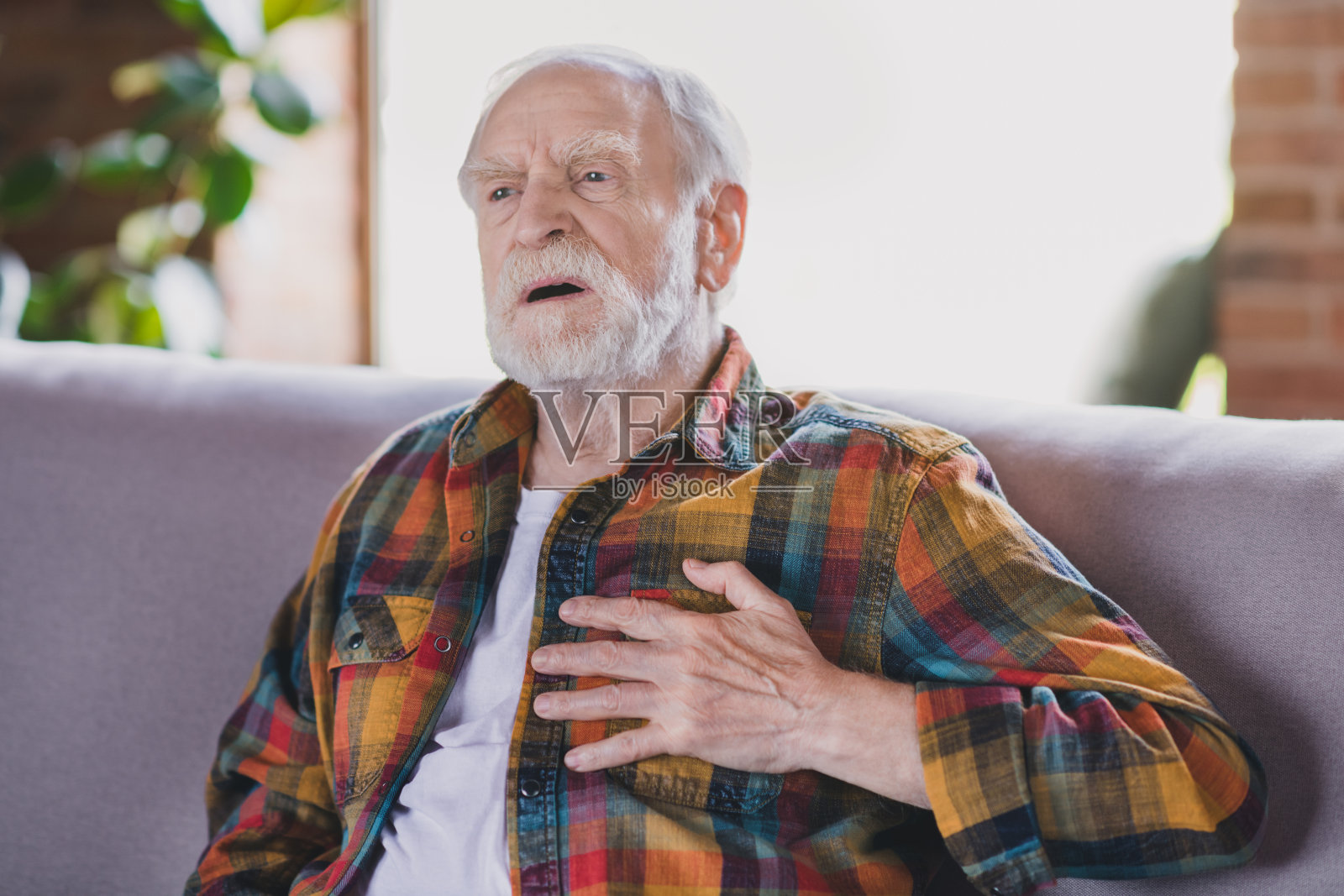 生病、压力大的爷爷，白发，手放在胸口，心脏问题，穿着休闲格子衣服，坐在家里的沙发上。照片摄影图片