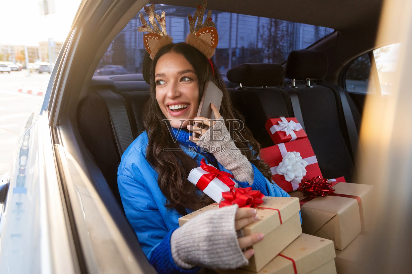 兴奋的女士坐在车里打电话，窗外望去面带微笑，车内有包裹好的礼物盒，正在为冬季假期准备和购买礼物。照片摄影图片