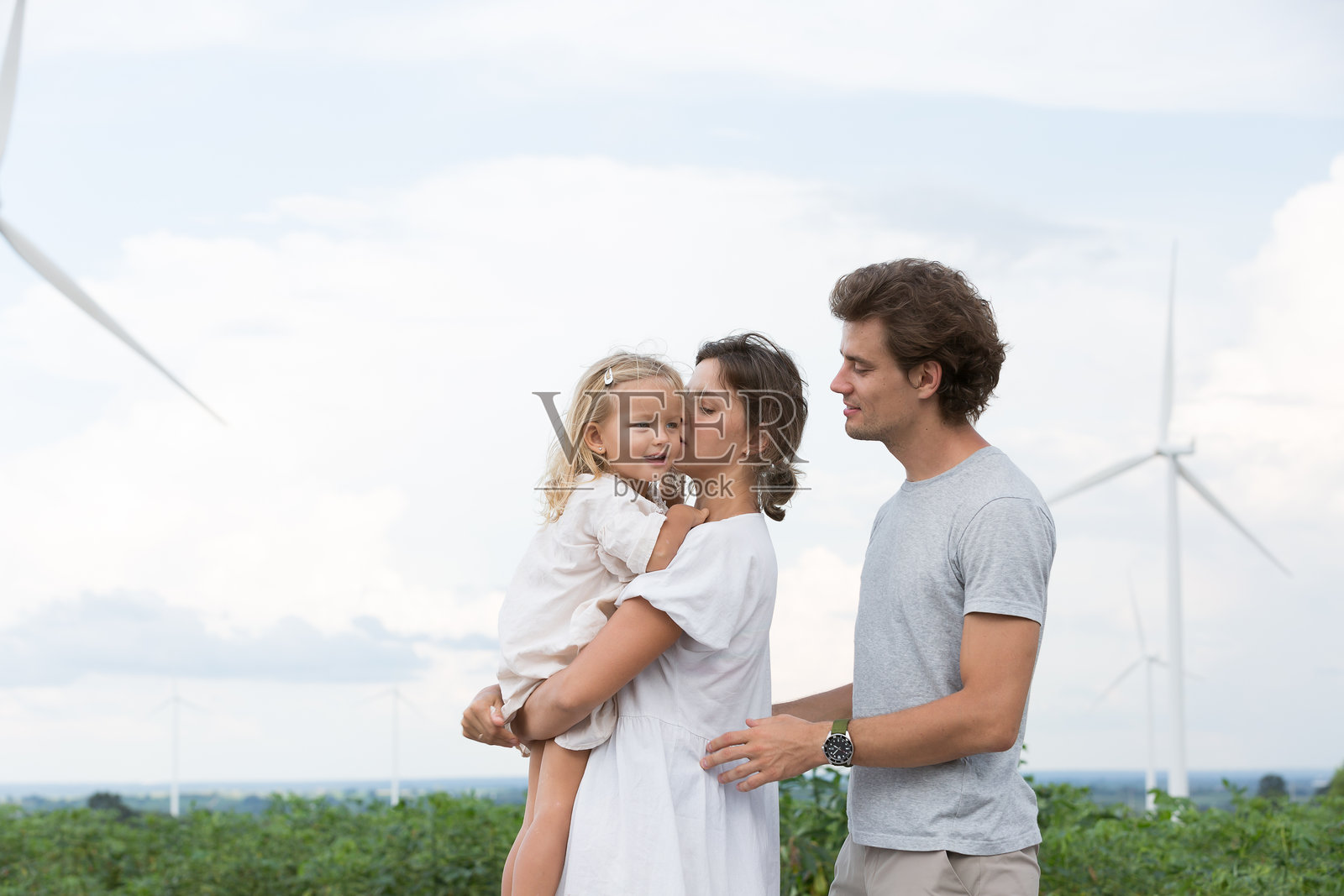 快乐的白人家庭在风力发电站度假旅行。母亲在风车田野中拥抱并抱着小女儿。小家庭旅行的概念。照片摄影图片